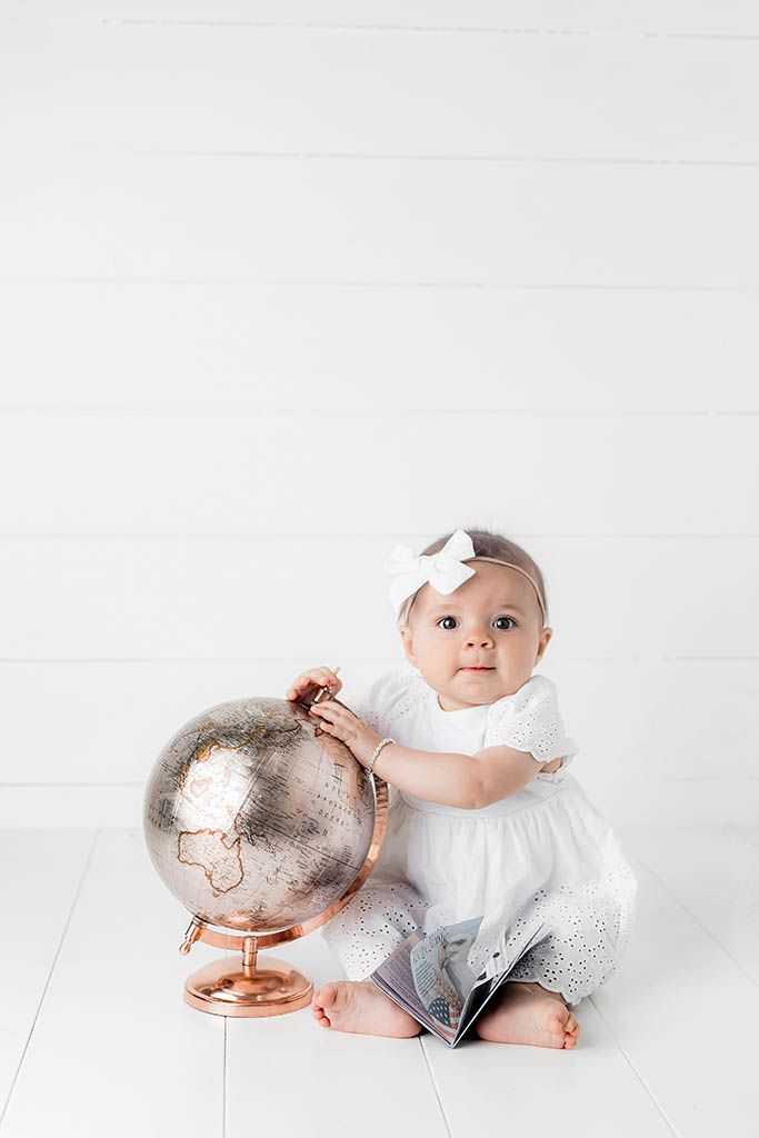 Baby girl in white eyelet outfit with bow headband sitting beside a rose gold globe.