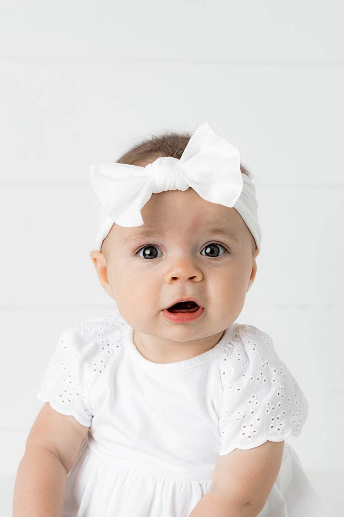 Cute baby girl wearing white eyelet dress and large white bow headband looking at camera.