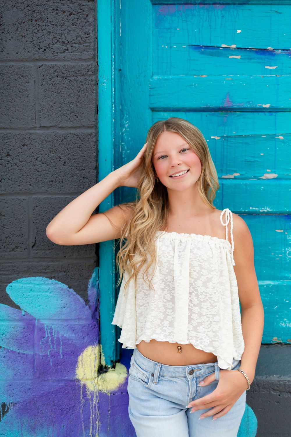 Smiling blonde teen in white lace crop top and jeans posing against colorful teal graffiti wall.