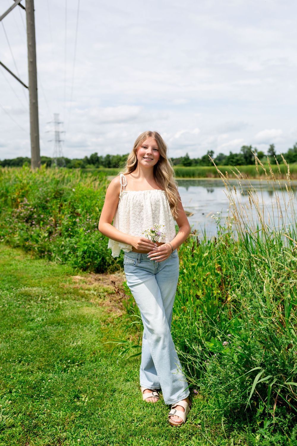 Young blonde woman in white top and jeans holding wildflowers by a serene pond on a sunny day.