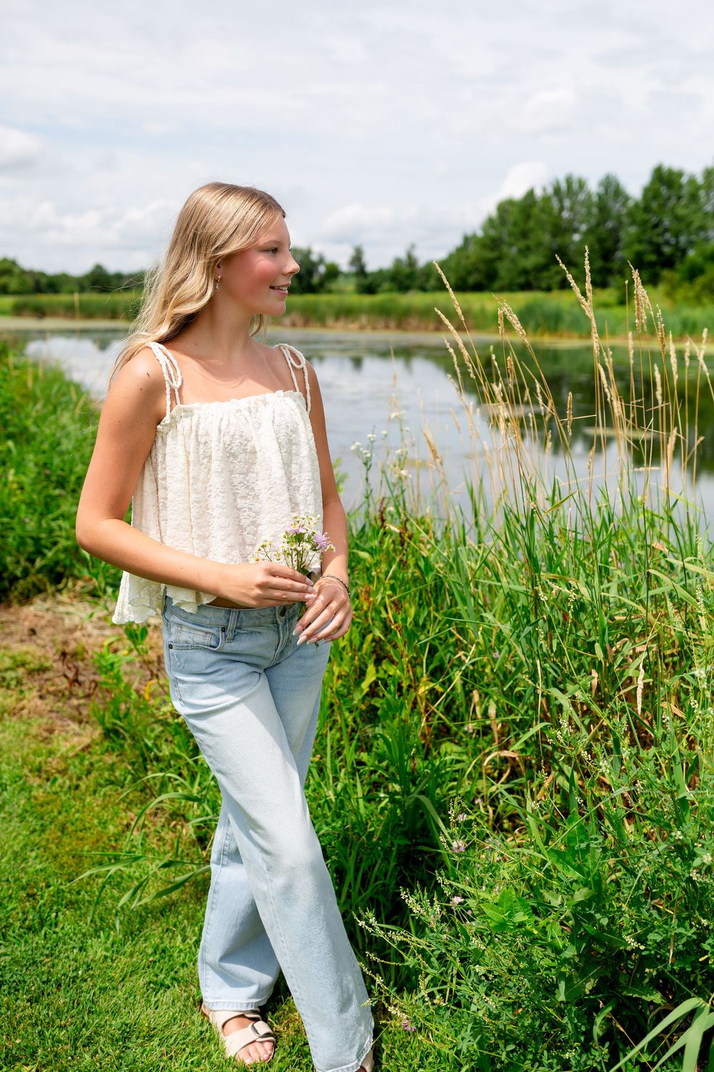 Young blonde woman in white top and jeans holding wildflowers by a serene pond surrounded by lush greenery.