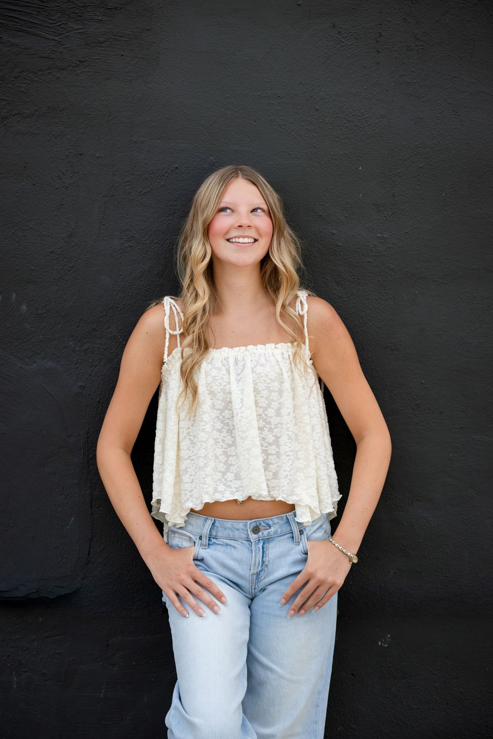Smiling blonde teen girl in white lace crop top and light blue jeans posing against black wall.