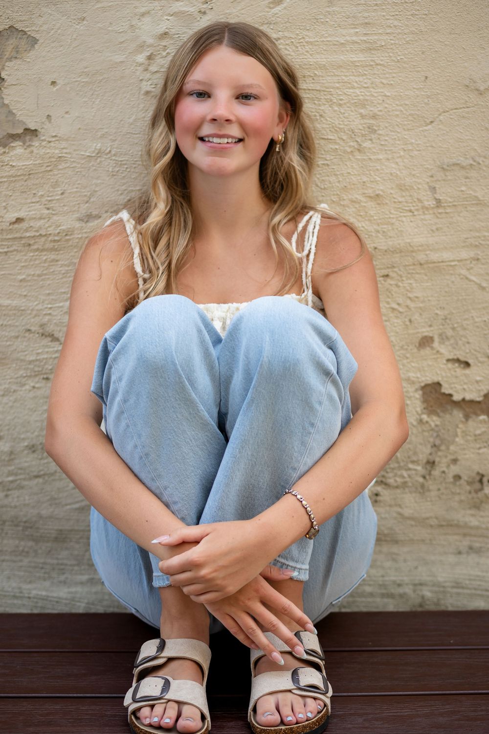 Smiling blonde teen girl in white lace top and jeans sitting cross-legged against a beige wall.