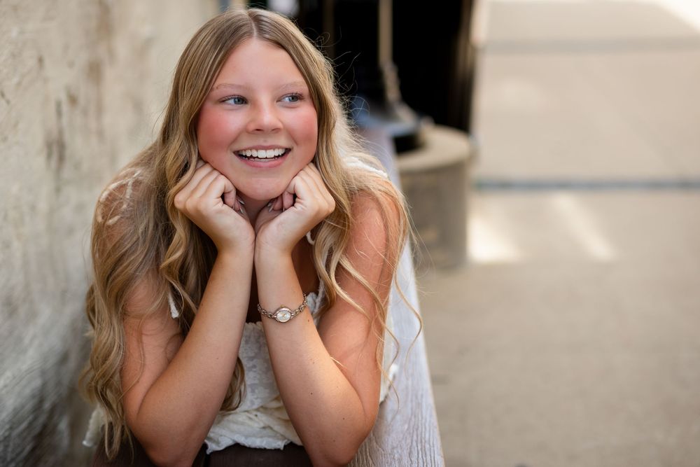 Smiling teenage girl with long blonde hair resting chin on hands, wearing a watch, posed outdoors.