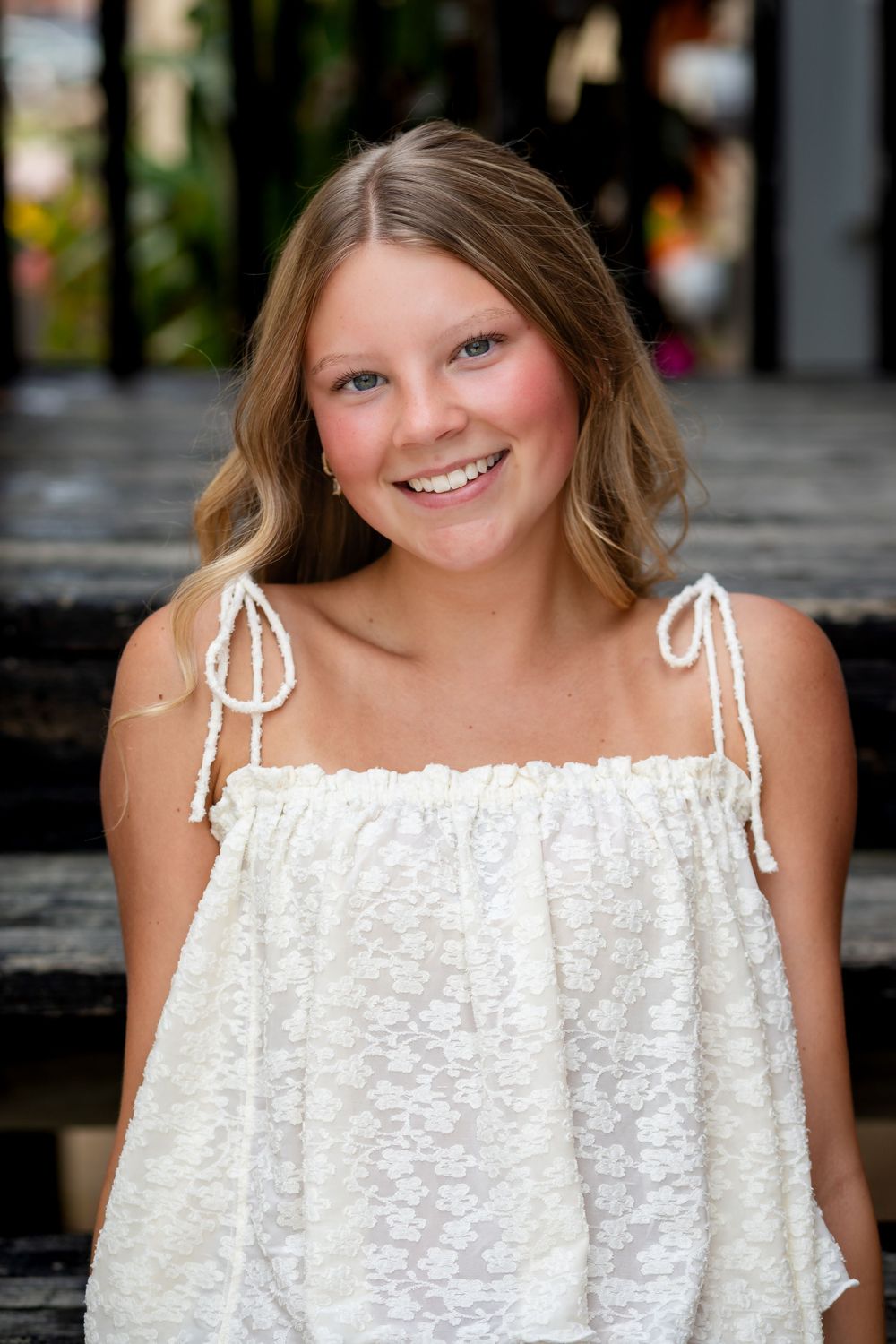 Smiling young woman with blonde hair wearing a white lace sundress, posed outdoors on wooden steps.