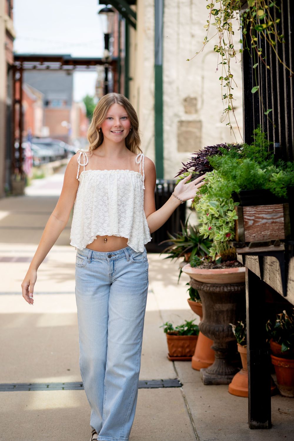 Smiling blonde teen girl in white lace crop top and wide-leg jeans walking on urban sidewalk.