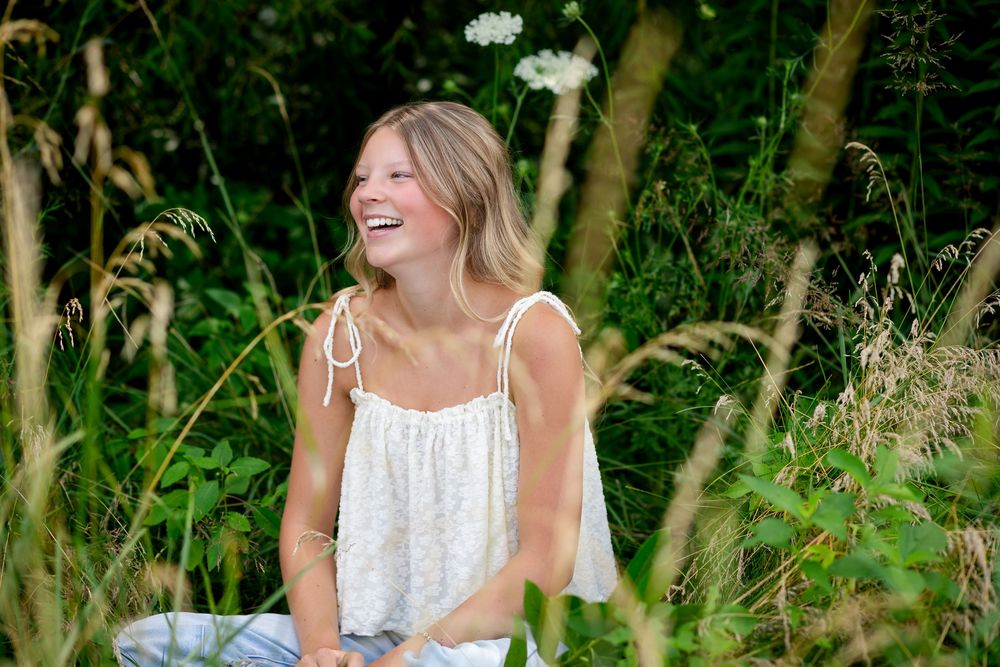 Young blonde woman laughing joyfully in a lush green meadow, wearing a white floral sundress.