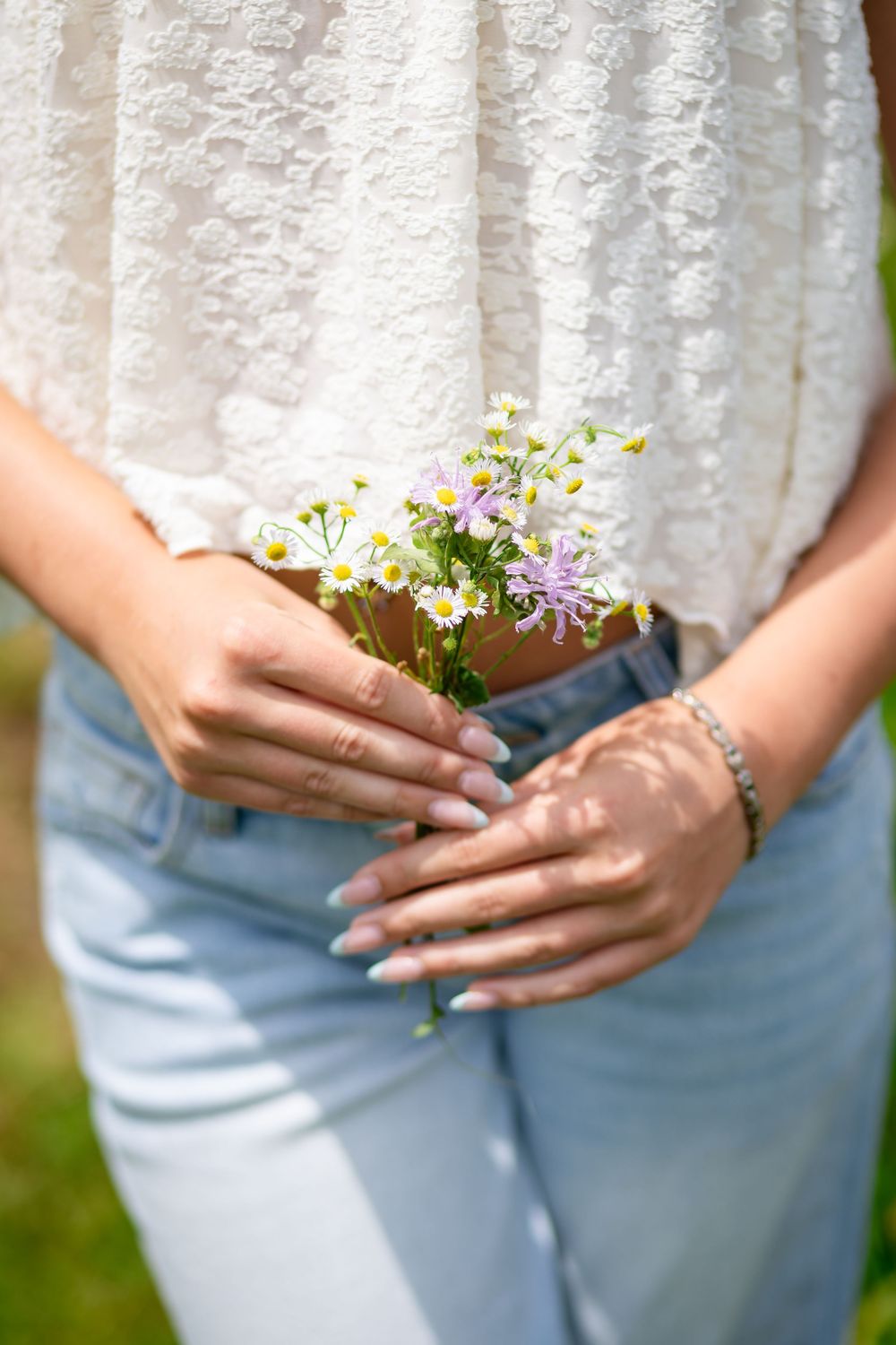Woman in white lace top and jeans holding a small bouquet of wild daisies and purple wildflowers in her hands.