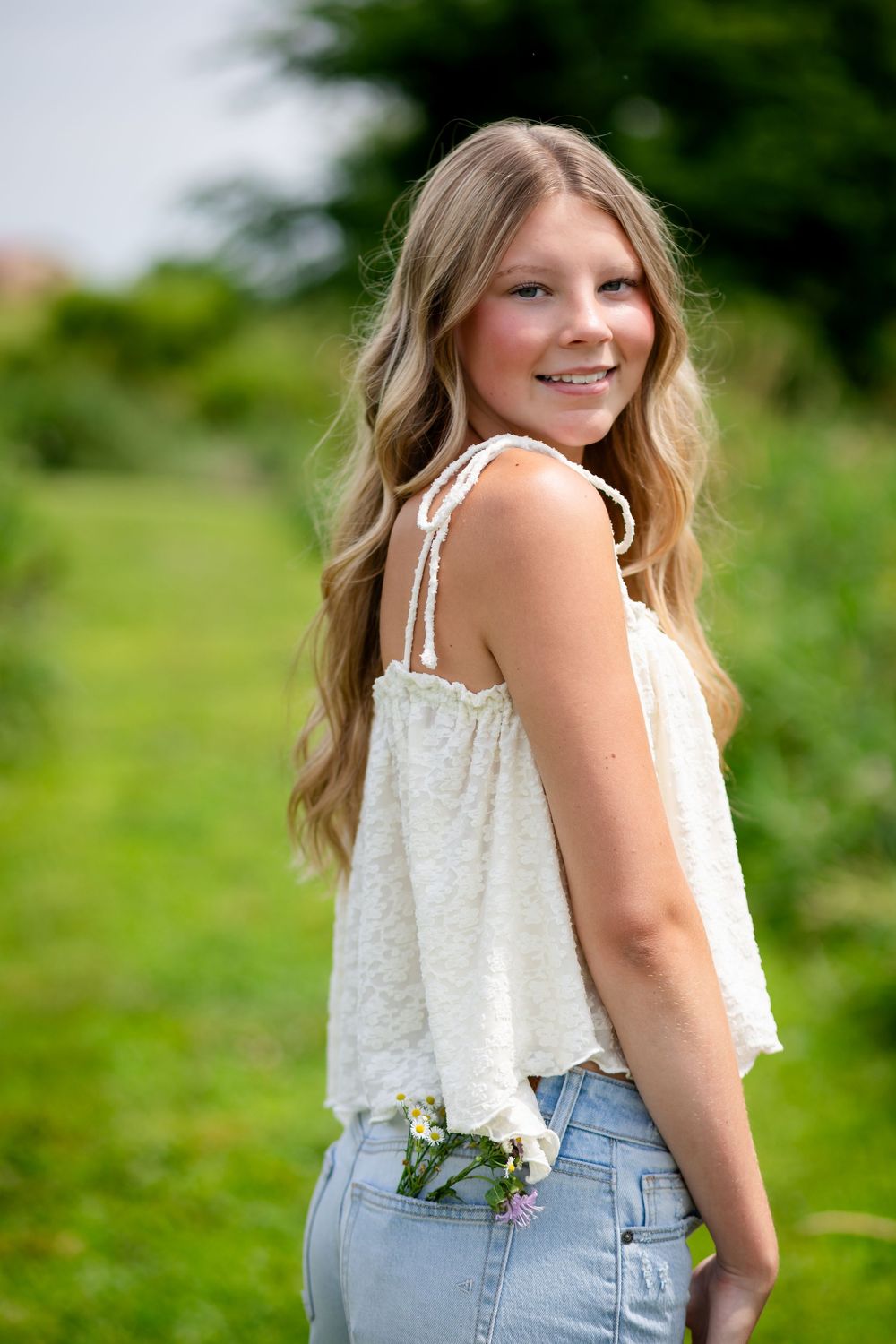 Young blonde woman in white lace top and jeans smiling over shoulder in green outdoor setting.