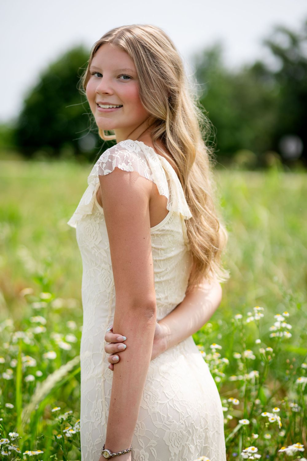Young girl in white lace dress smiling over shoulder in sunny wildflower meadow with daisies and green trees.