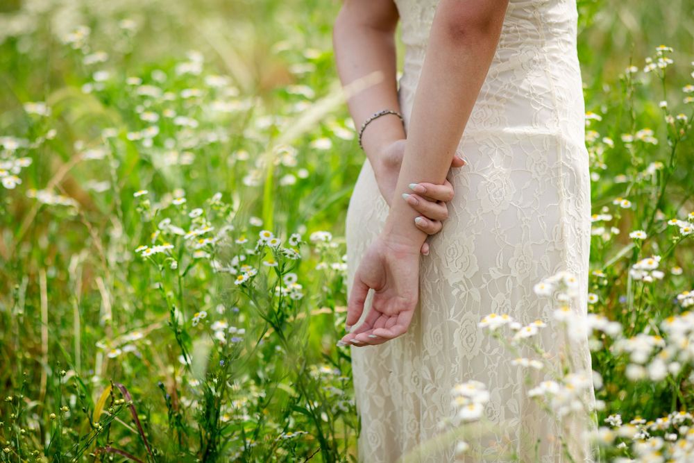 Bride in white lace dress walking through a wildflower meadow, hands gently touching blooming daisies.