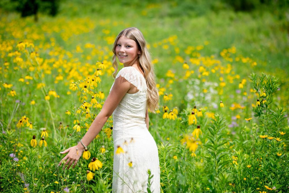 Young blonde woman in white lace dress posing in a vibrant yellow wildflower meadow, looking back over her shoulder.