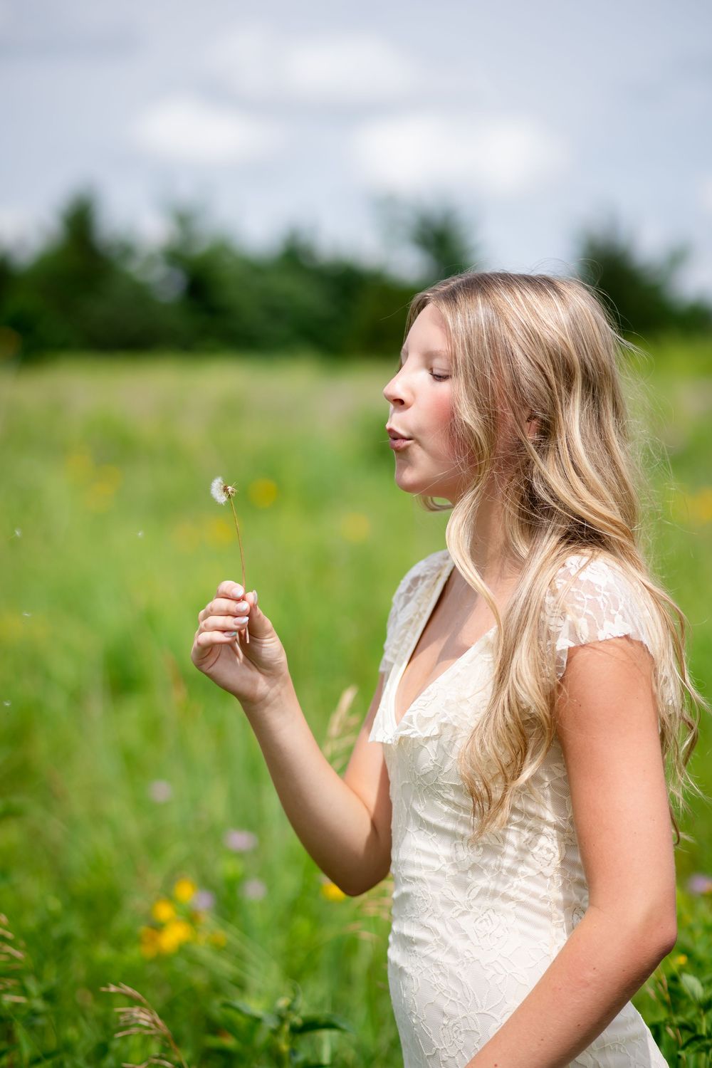 Young blonde woman in white lace dress blowing dandelion seeds in a green meadow on a cloudy day.