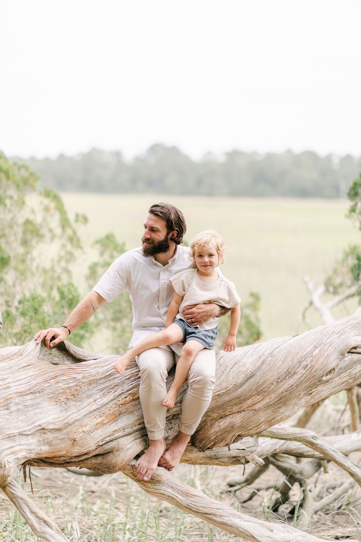 Bearded father and curly-haired toddler sitting on a fallen tree trunk in a scenic green field outdoors.