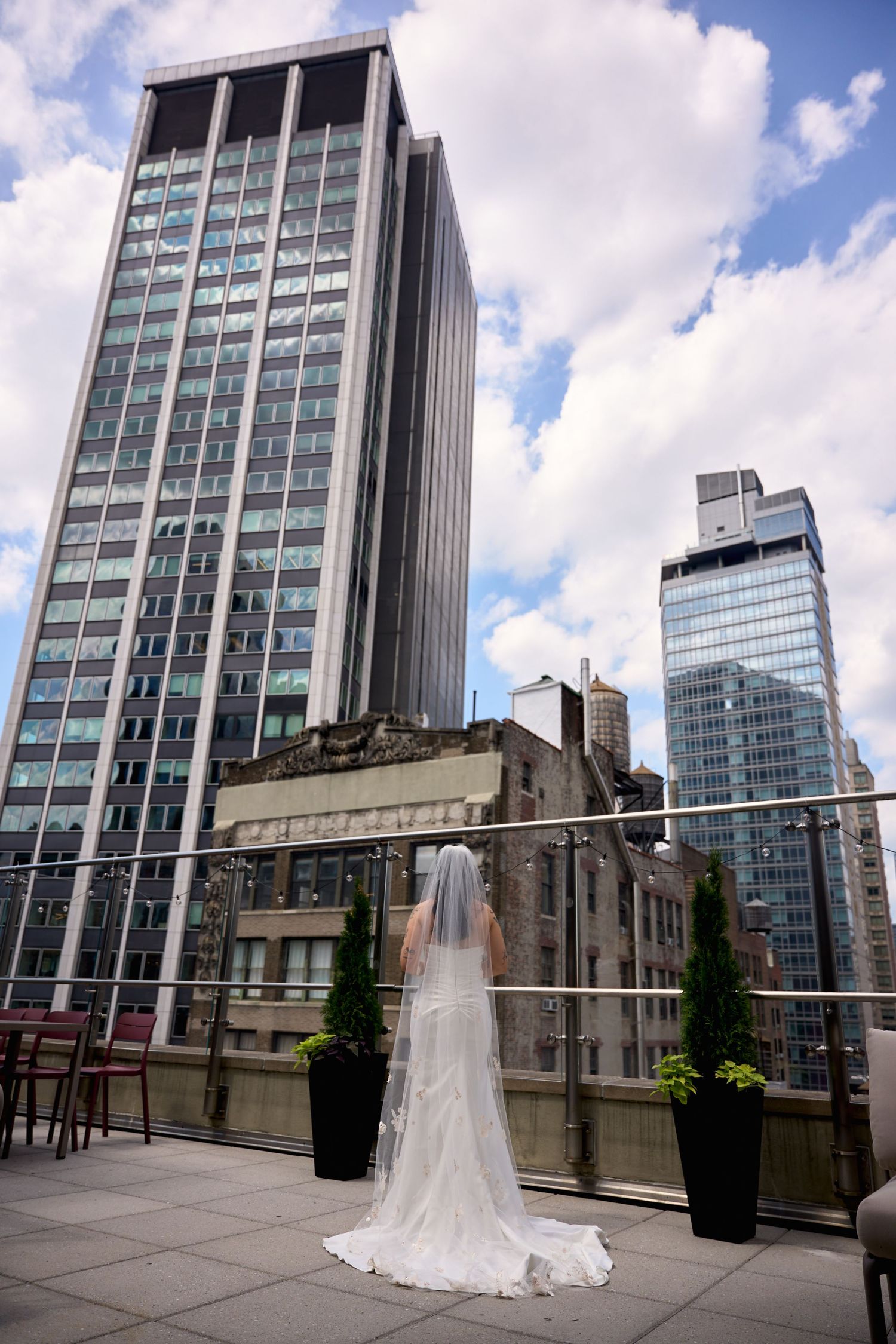Bride in white gown and veil stands on rooftop terrace overlooking NYC skyline on cloudy day.