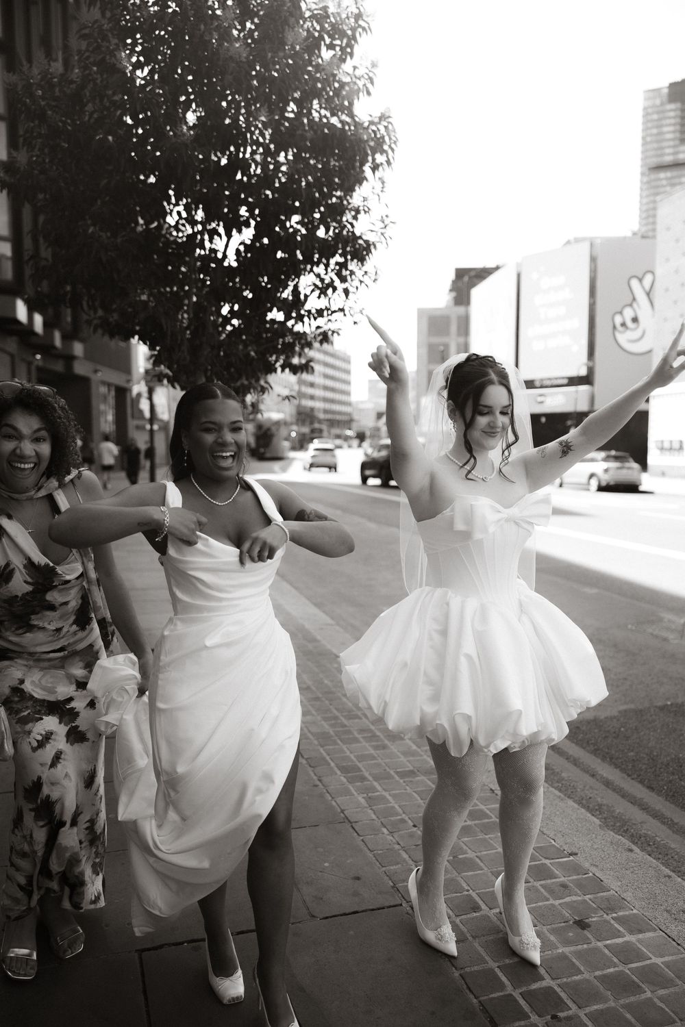 Bride and bridesmaids dancing joyfully on a city sidewalk in black and white wedding photo.