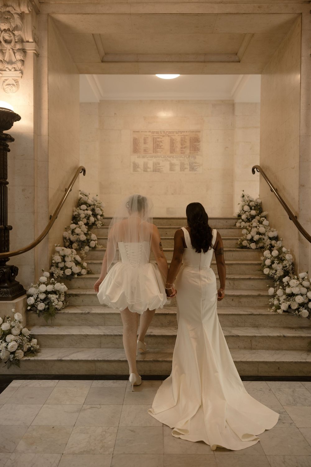 Two brides in white gowns hold hands ascending floral-adorned marble stairs in an elegant wedding venue.