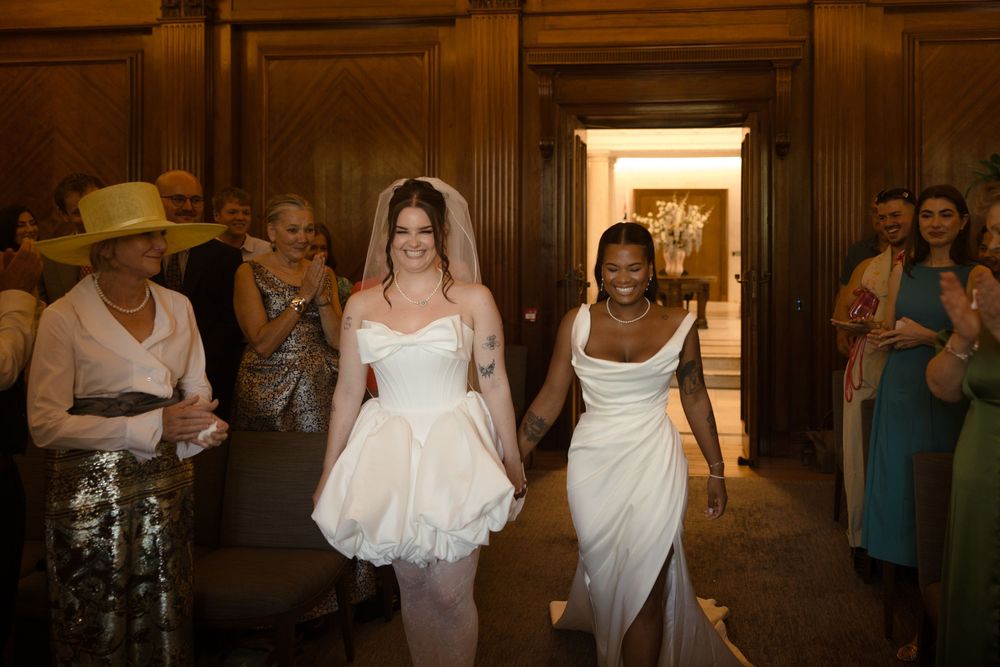 Two brides in white wedding gowns walk hand in hand down the aisle surrounded by smiling guests.