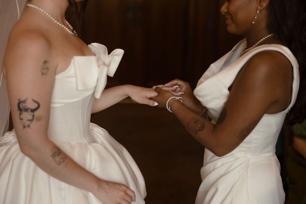 Two brides in white gowns exchange rings during an intimate wedding ceremony, both wearing pearl necklaces.