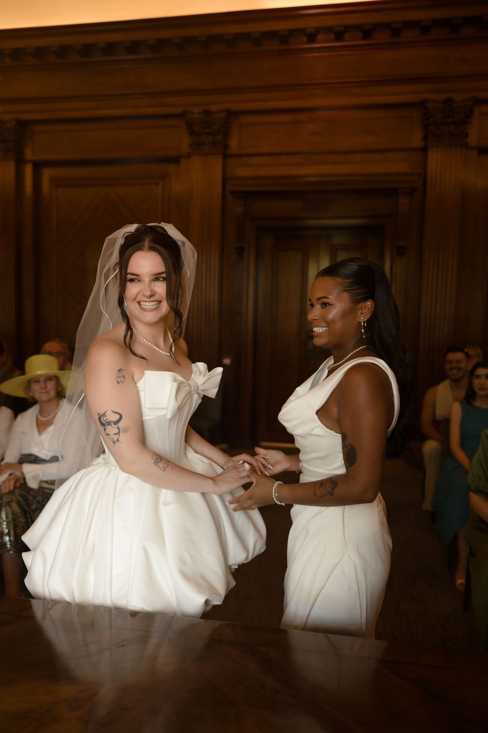 Two brides in white dresses hold hands and smile during their wedding ceremony in an elegant wood-paneled venue.