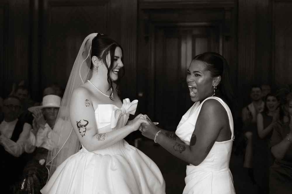 Two brides exchange rings during wedding ceremony, one in veil and gown, other in white dress, guests applauding in background.