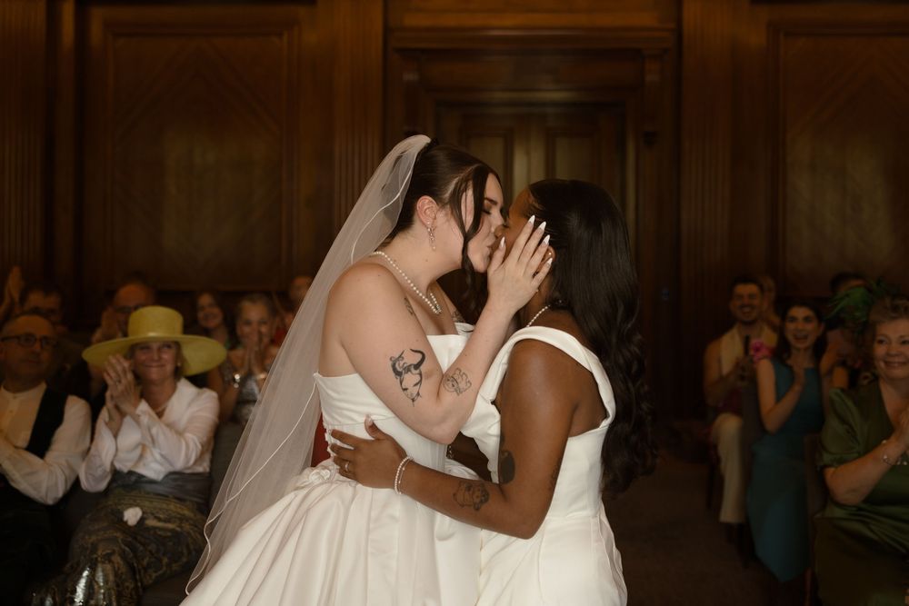 Two brides sharing a first kiss at their wedding ceremony, surrounded by applauding guests.