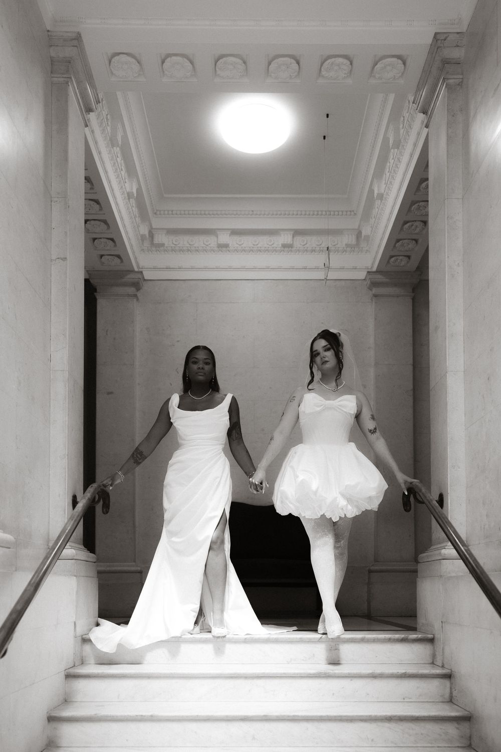 Two brides in white wedding gowns hold hands descending marble stairs in an elegant venue, black and white photo.