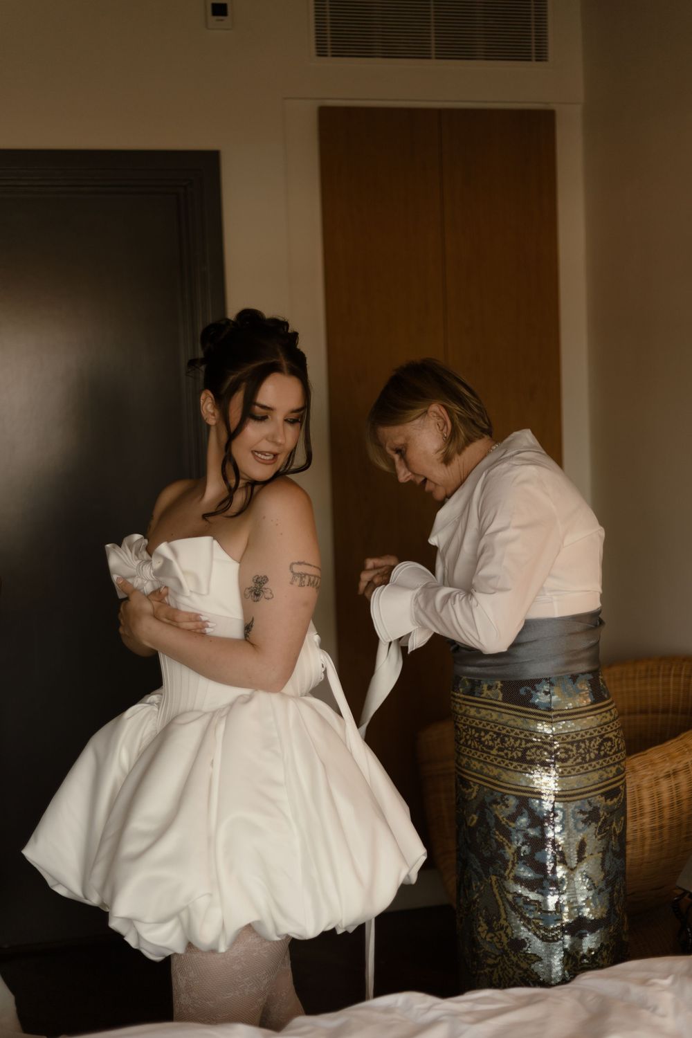 Bride in short white puffball dress being helped by assistant in hotel room before wedding ceremony.