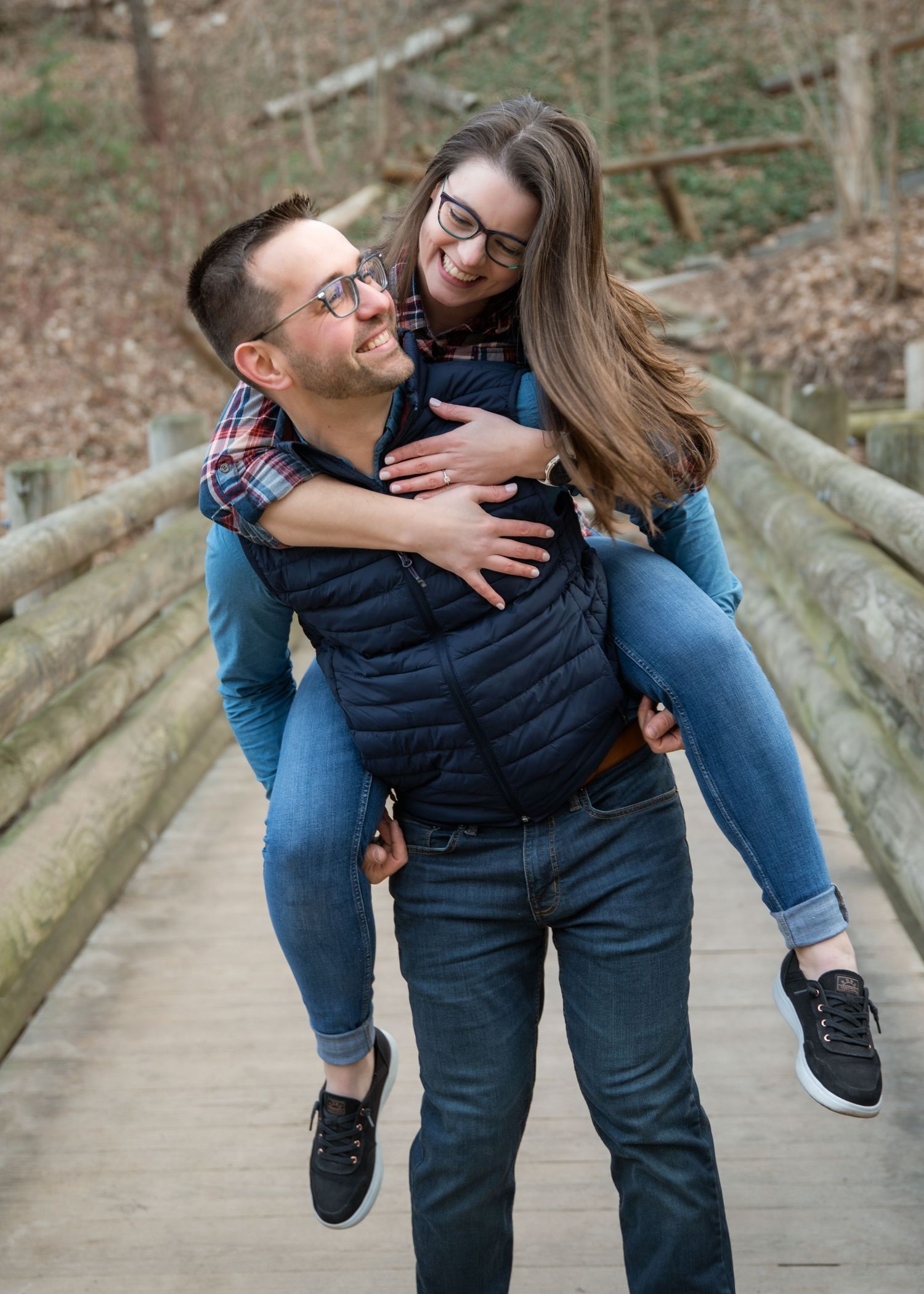 A man gives a woman a piggyback ride on a wooden bridge, both smiling and wearing glasses and casual blue outfits.