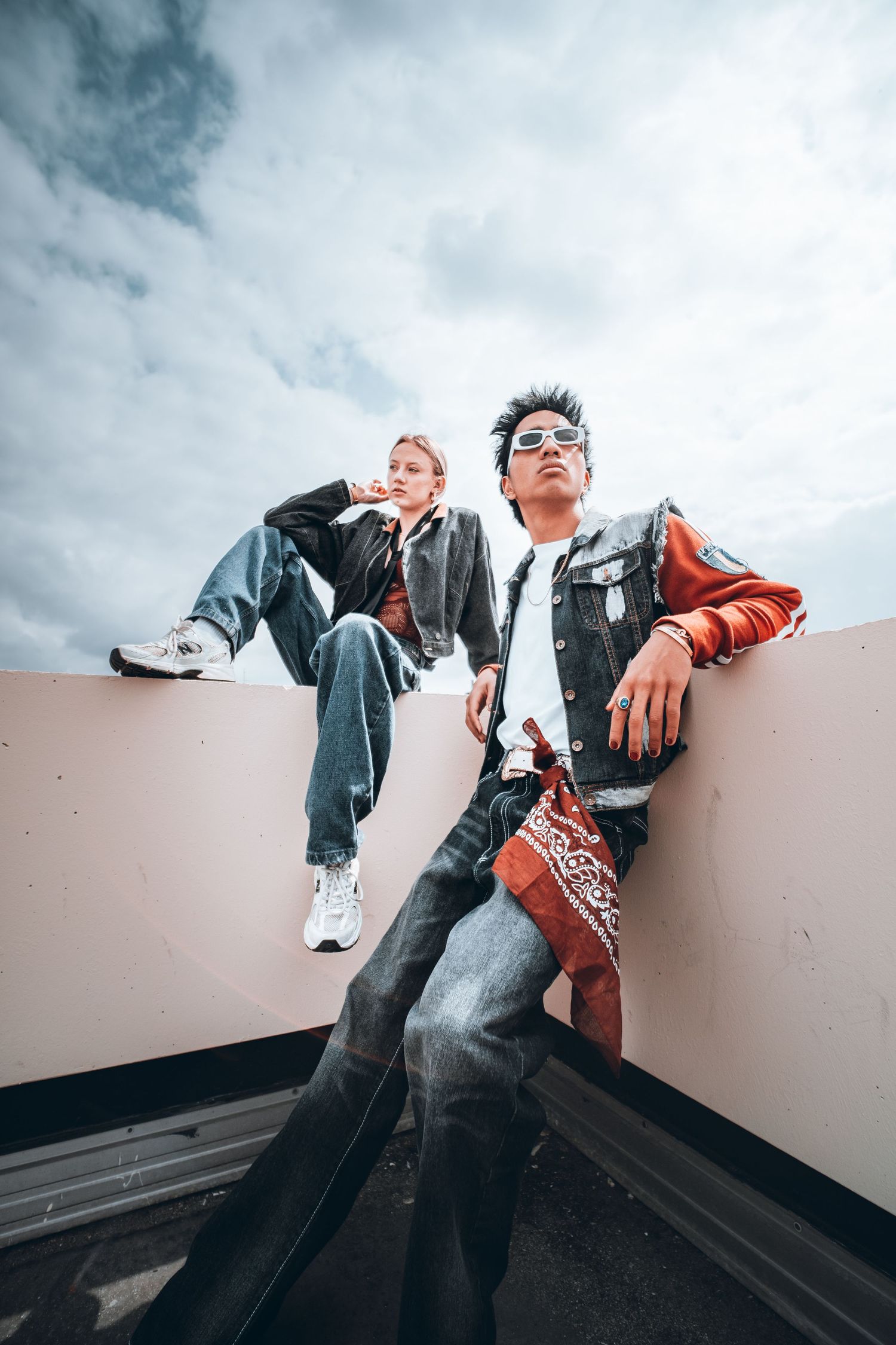 Two stylish young people pose on a rooftop ledge wearing denim jackets and jeans against a cloudy sky.