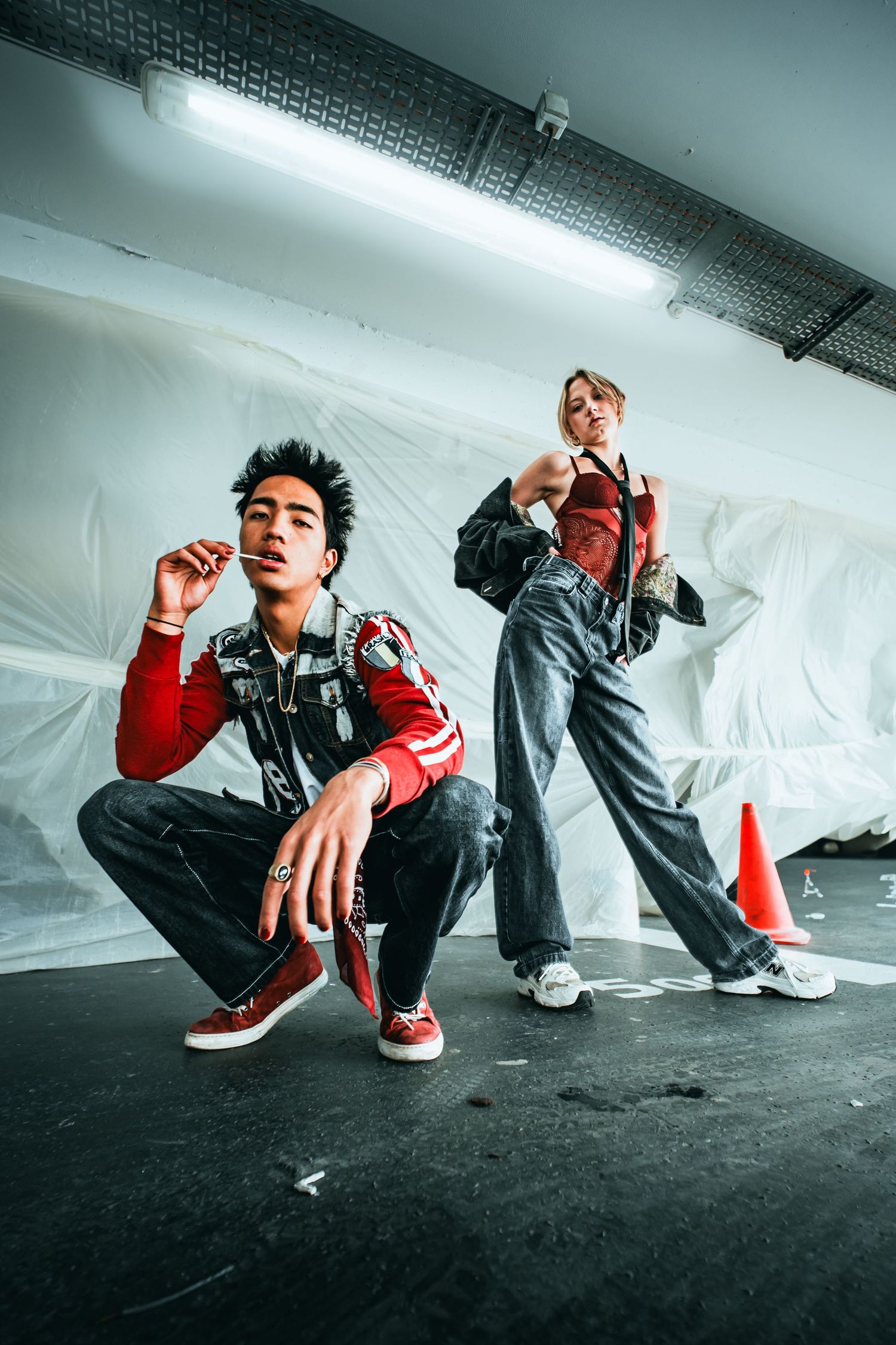 Two stylish young people pose in an urban setting, wearing red and gray streetwear outfits under fluorescent lights.
