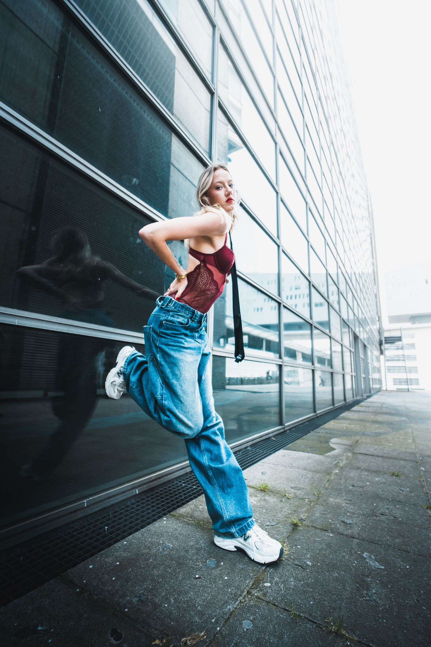A young woman in blue jeans and red top poses dynamically on a urban sidewalk beside a modern glass building.