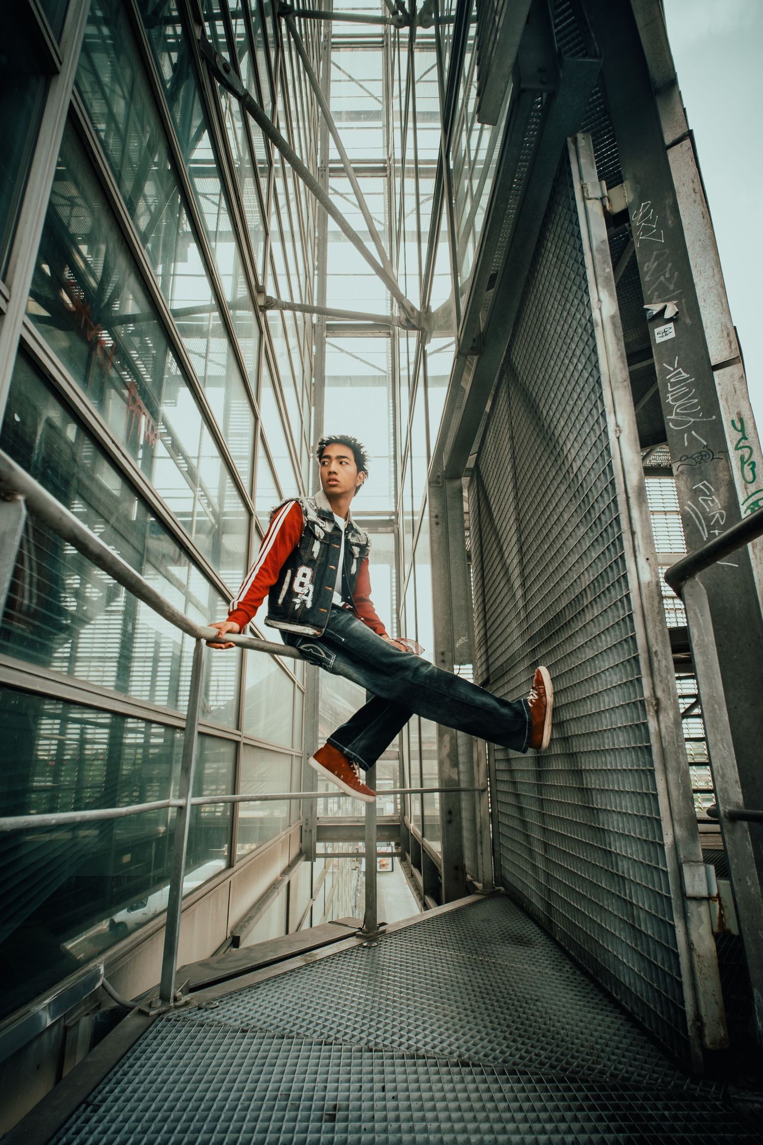 Young man in red jacket and jeans balancing on metal railings outside a modern glass building.
