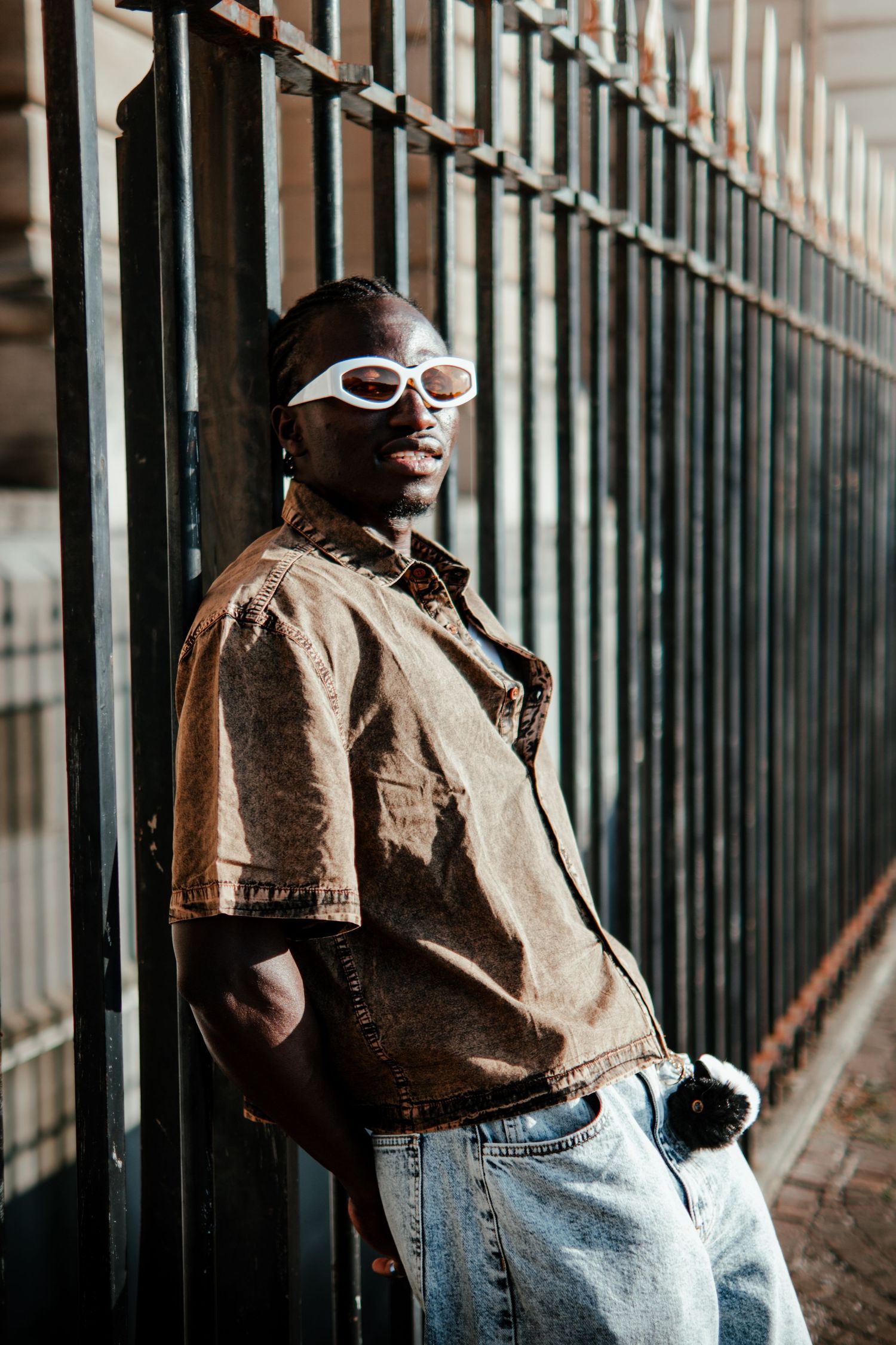A young Black man in a brown denim shirt, white sunglasses, and jeans leans against a metal fence outdoors.
