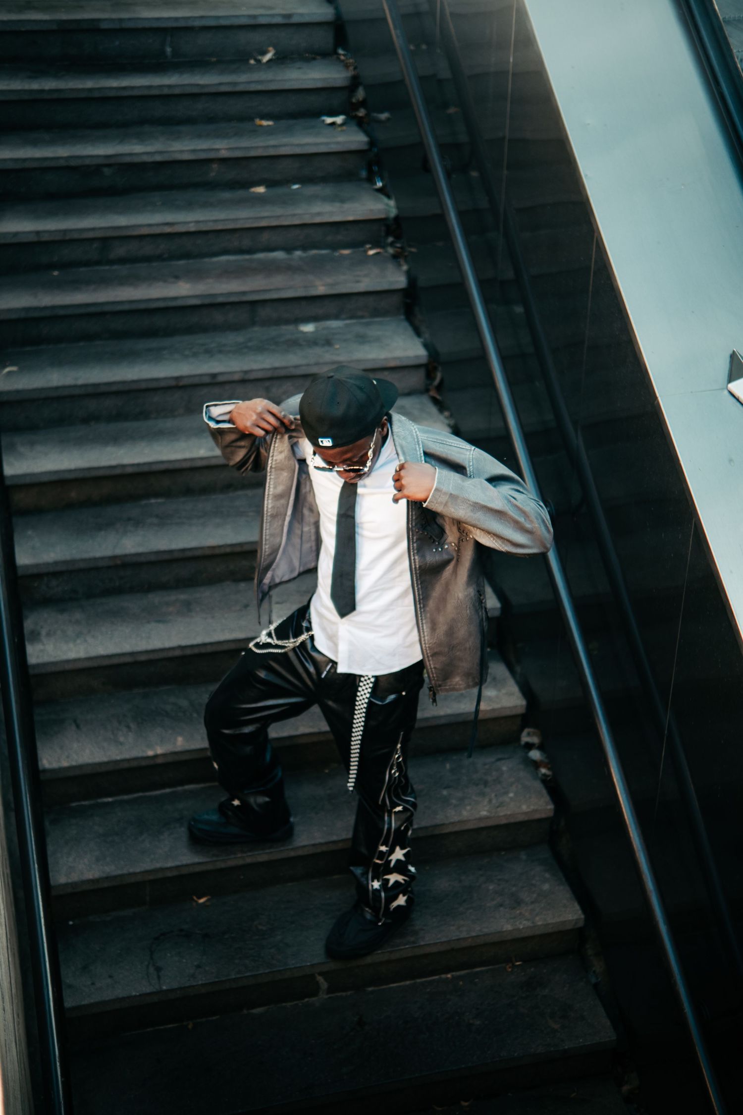 Stylish man in cap, blazer, and star-print pants posing on escalator stairs in an urban setting.