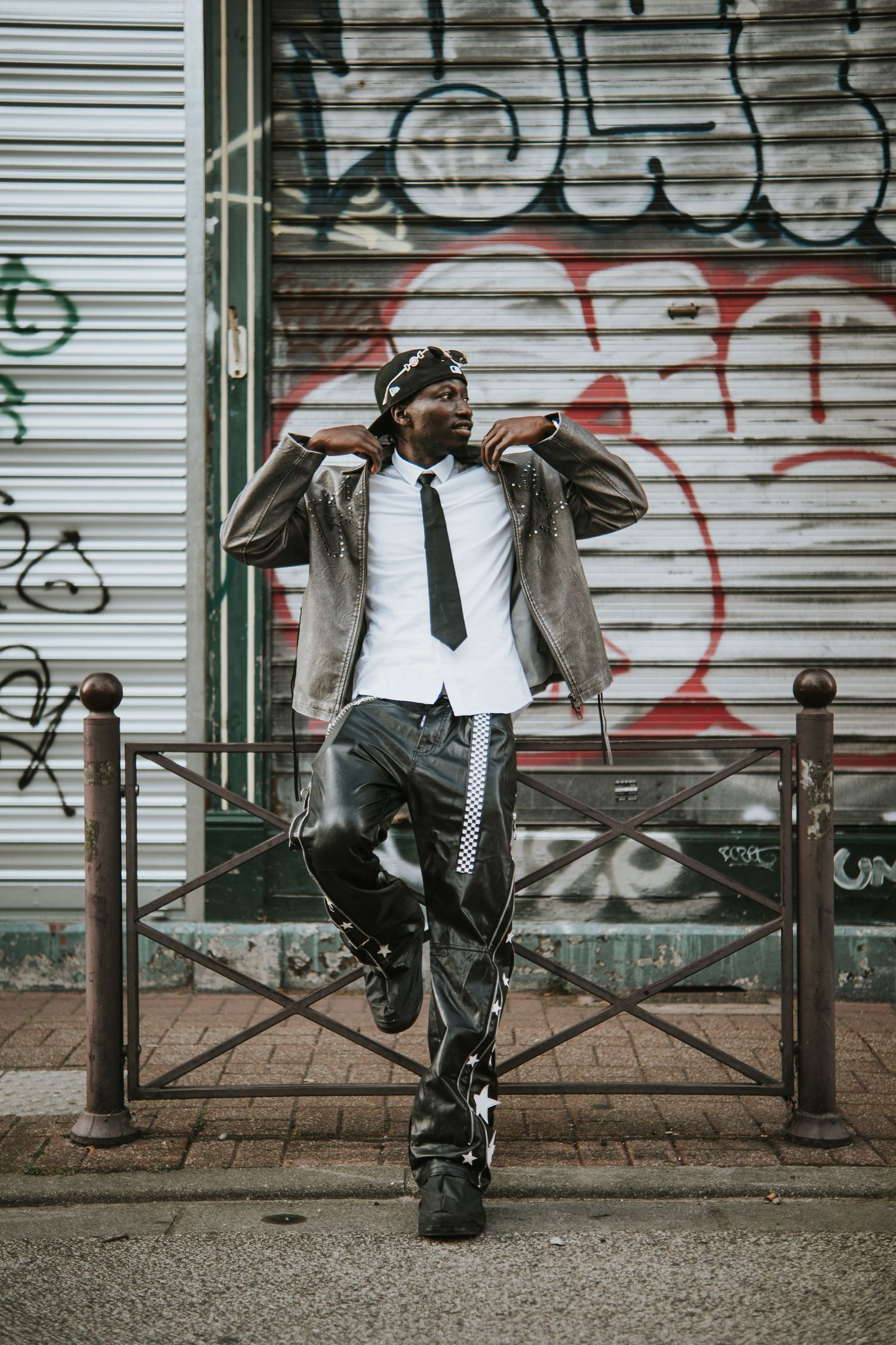 Stylish Black man posing in urban street wear with tie, leather pants, and cap against graffiti wall.