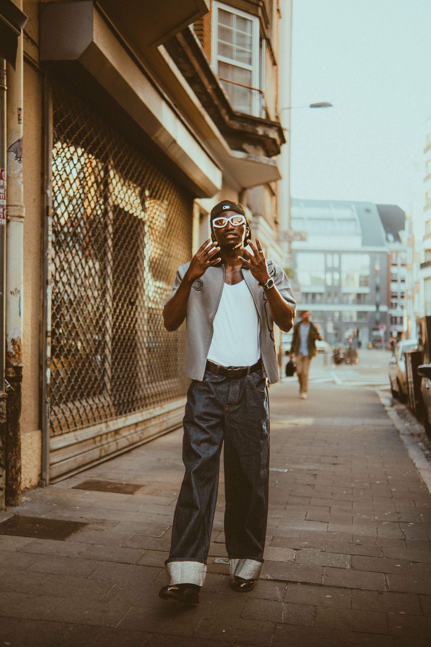 A stylish man poses on a urban sidewalk wearing wide-leg pants, white tank, and bold sunglasses in warm sunlight.