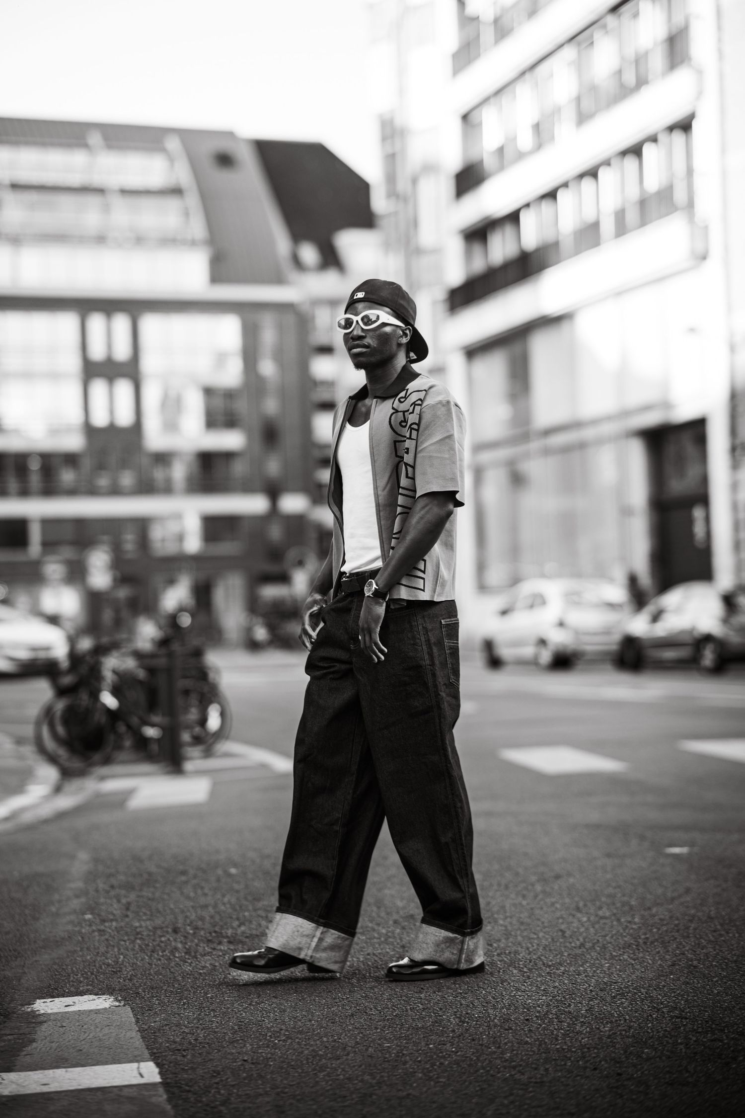 Black and white street photo of a stylish man in baggy jeans, cap, and sunglasses crossing an urban street.