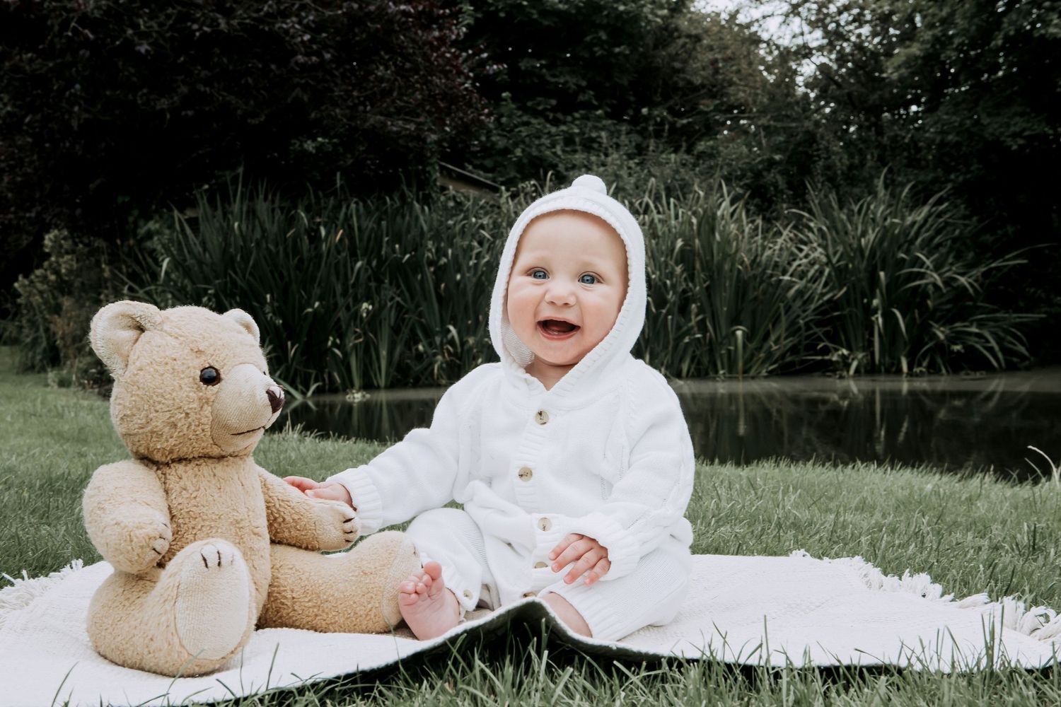 Smiling baby sitting beside a teddy bear during an outdoor baby photography session in Bristol.