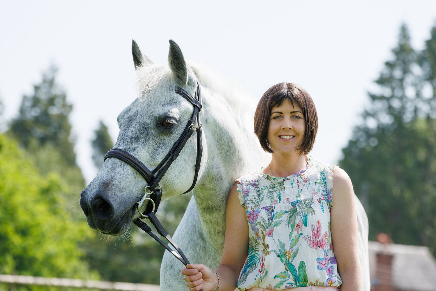 A woman sits on a log beside a brown horse in a field, holding its reins in a serene black and white photo.