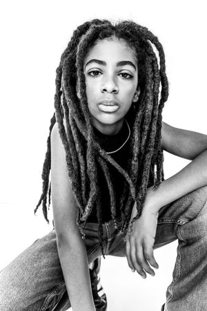 Black and white portrait of a young person with long dreadlocks wearing a sleeveless top and jeans.