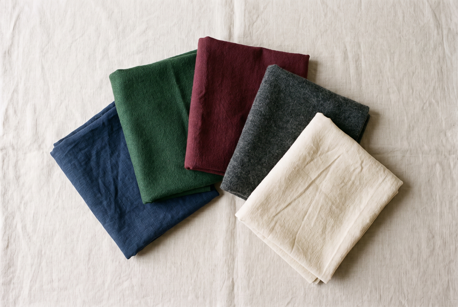 Five folded linen napkins in navy, green, burgundy, charcoal, and cream colors arranged in a fan display.