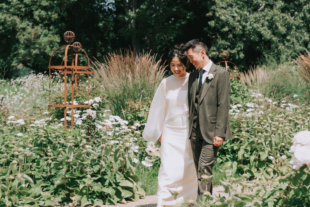 Bride and groom walking through a lush garden on their wedding day, surrounded by wildflowers and greenery.