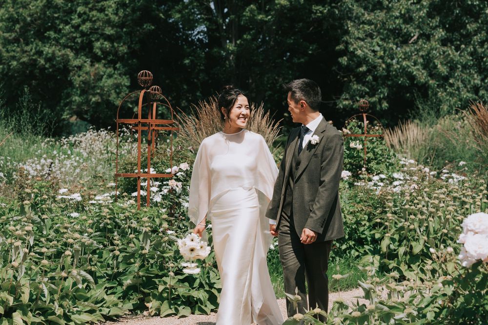 Asian bride in white cape dress and groom in grey suit walk through a lush garden on their wedding day.