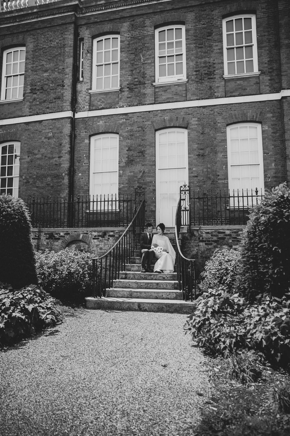 Black and white photo of wedding couple sitting on steps outside elegant brick building with iron railings.