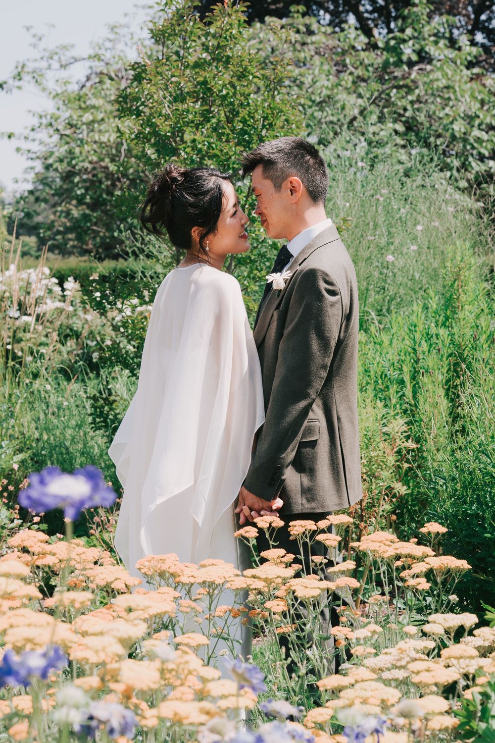 Bride and groom holding hands facing each other surrounded by wildflowers in lush garden.
