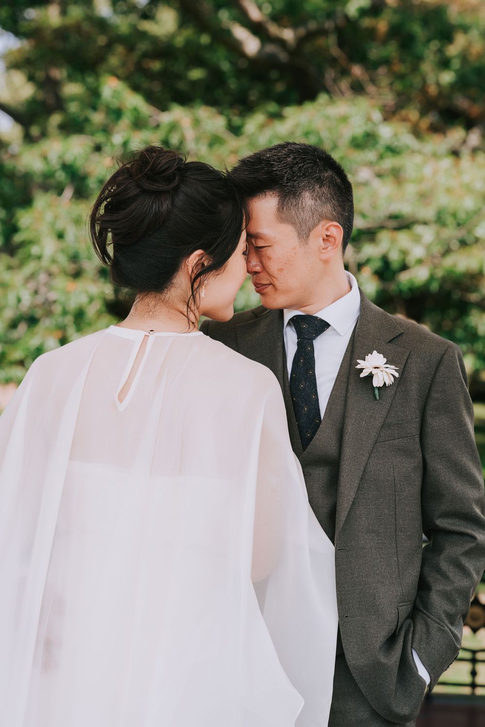 Asian groom in grey three-piece suit with floral boutonniere shares a tender forehead touch with his bride outdoors.