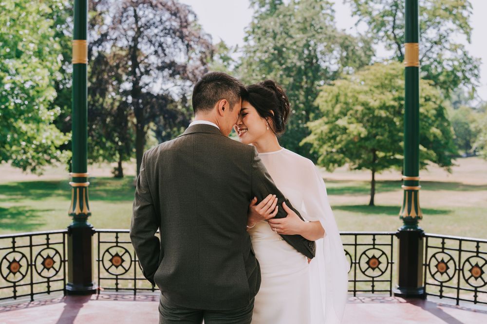 A couple in wedding attire shares a romantic moment under a green gazebo in a lush park setting.