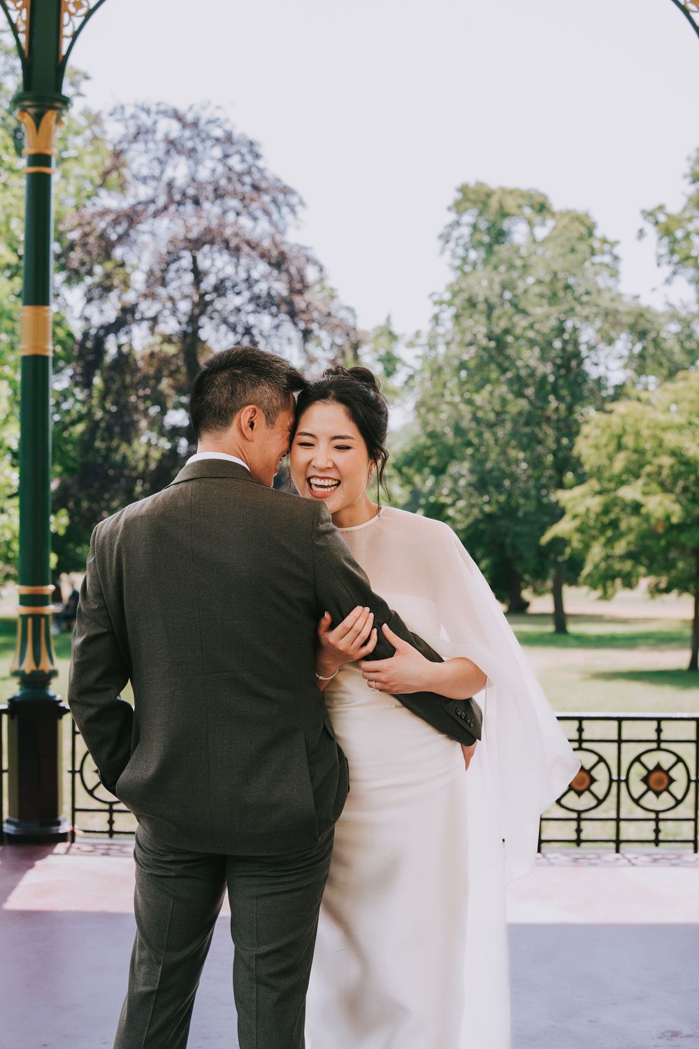 Bride in white cape gown laughs with groom in gray suit at an outdoor park wedding ceremony.