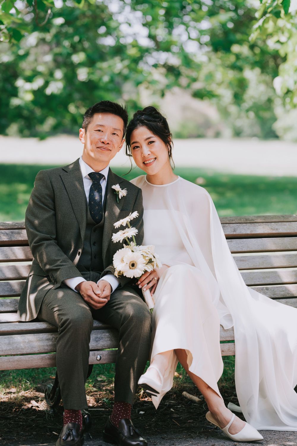Asian couple in wedding attire sitting on park bench, bride in white gown holding bouquet, groom in grey suit.